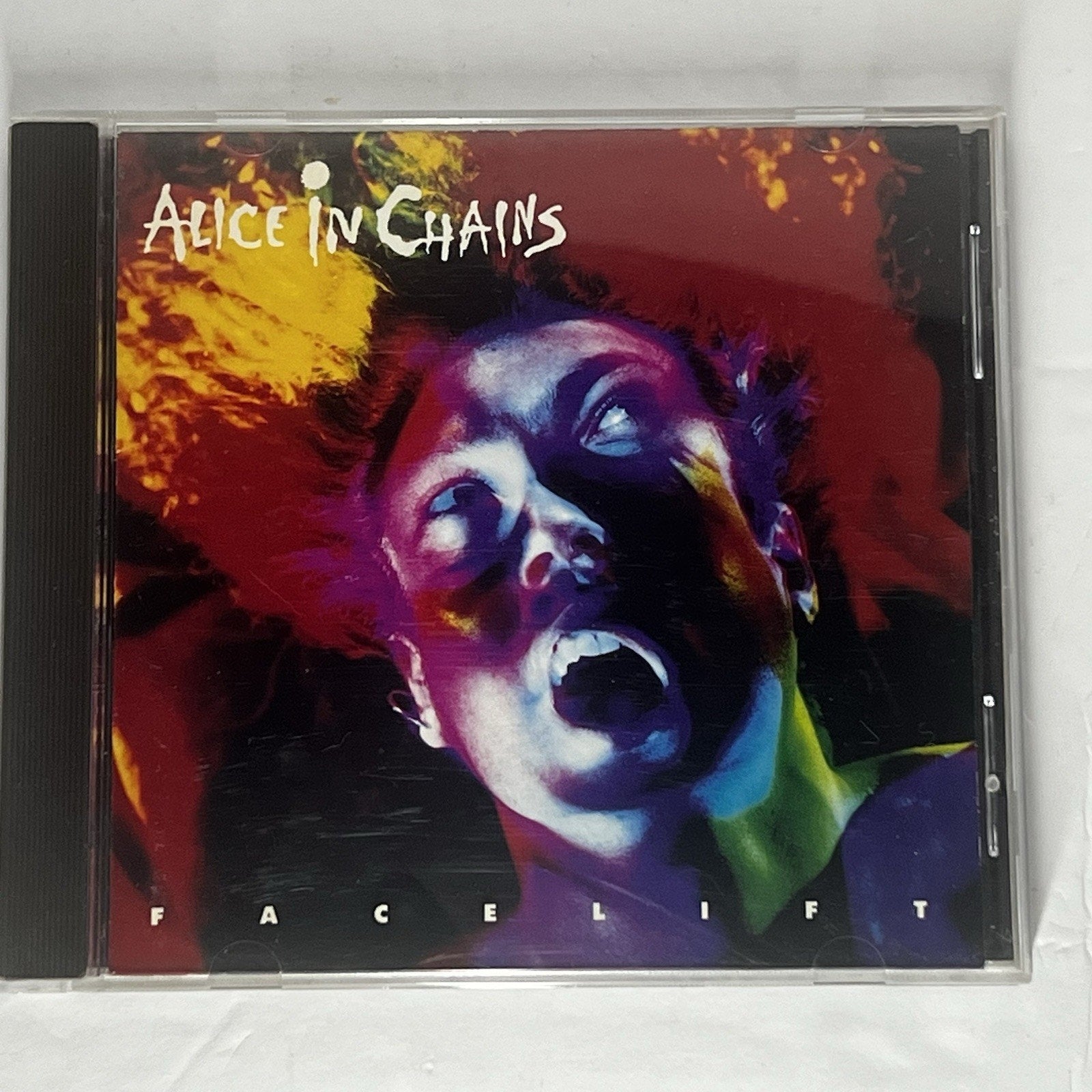 Alice in Chains 'Facelift' CD album in a clear jewel case, photographed from above on a white fabric background. The cover art features a dramatic close-up portrait with bold red, purple, yellow, and blue color blocking.