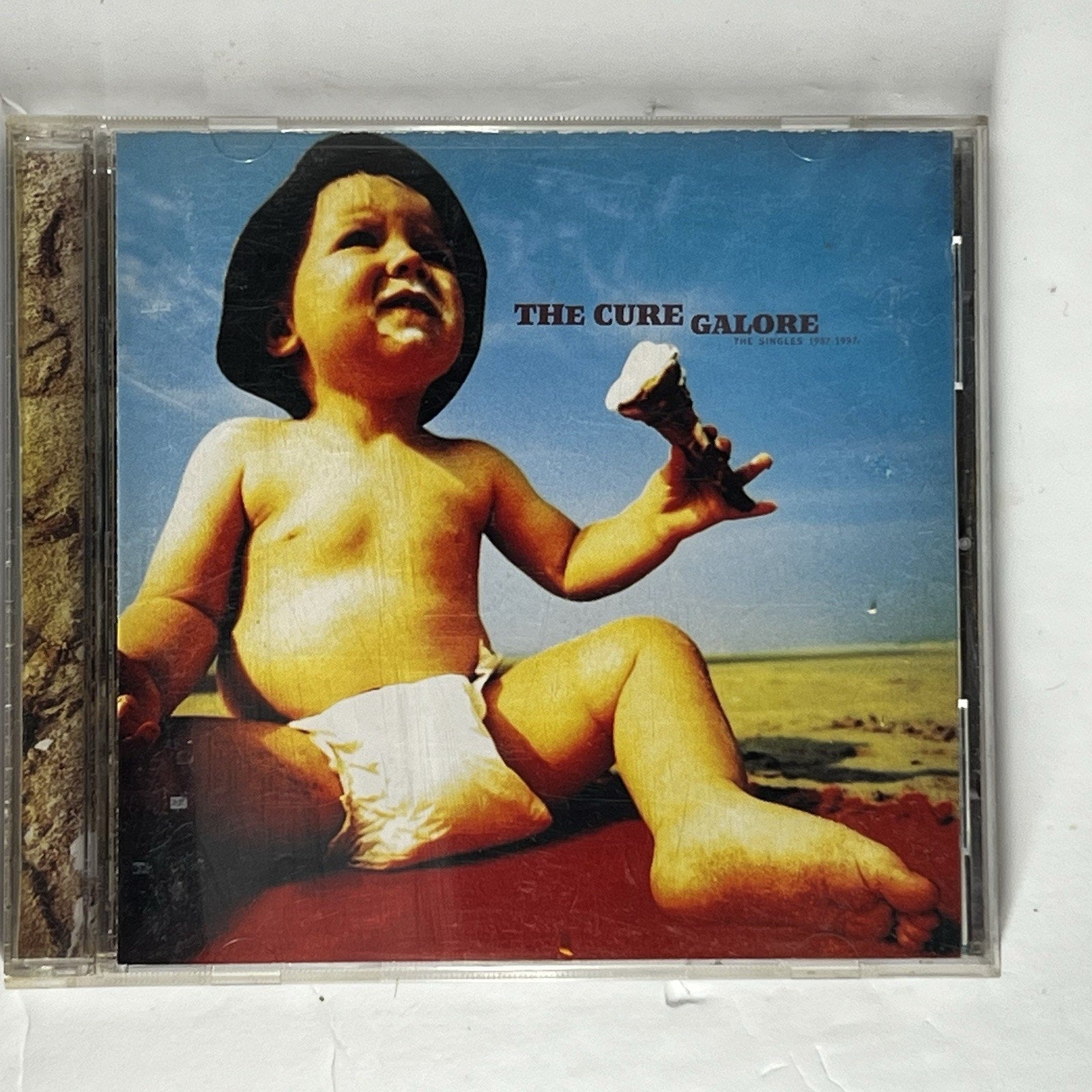 CD album case for The Cure's 'Galore' featuring the album cover art of a baby sitting outdoors under a blue sky.