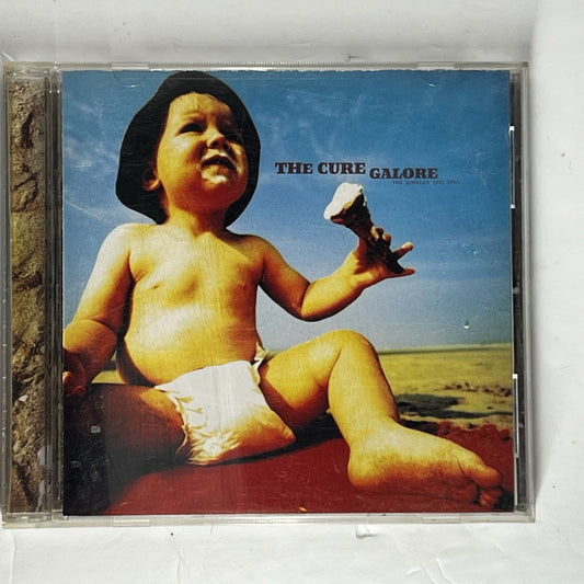 CD album case for The Cure's 'Galore' featuring the album cover art of a baby sitting outdoors under a blue sky.